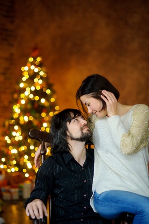 Christmas evening. Young woman embraces man sitting in a chair. She sits on an armrest. Young people with love look at each other. In a room corner stands Christmas tree. She shines in the twilight.の写真素材