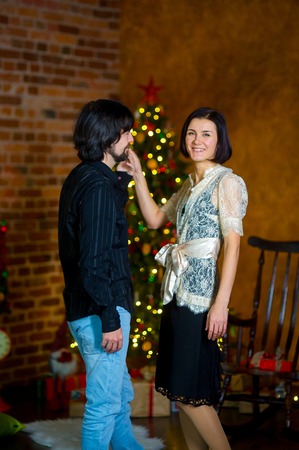 Young nice man invites woman to dance. Elegantly dressed woman smiles. In a corner of the room the Christmas tree decorated with garlands shines. Under her there are a lot of gifts in beautiful boxes.の写真素材