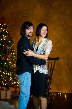 Nice young couple stands near the Christmas tree. The young man gently embraces the girlfriend. The woman is elegantly dressed. The Christmas tree is festively decorated.の写真素材