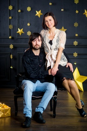 Young woman embraces man sitting in chair. She sits on an armrest. Dark wall of the room is decorated with gold garlands. On floor box with gift in gold packing lies. Young people look in the camera.の写真素材