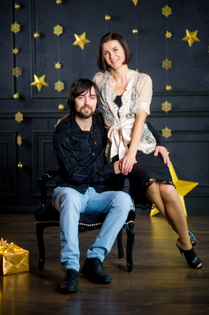 Young woman embraces man sitting in chair. She sits on an armrest. Dark wall of the room is decorated with gold garlands. On floor box with gift in gold packing lies. Young people look in the camera.の写真素材