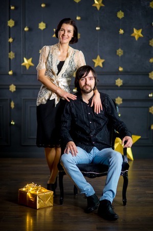 Young woman embraces man sitting in a chair. The dark wall of the room is decorated with gold garlands. On a floor the box with a gift in gold packing lies. Young people look in the camera.の写真素材