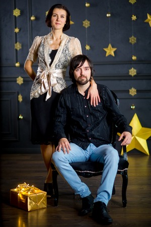 Young woman embraces man sitting in a chair. The dark wall of the room is decorated with gold garlands. On a floor the box with a gift in gold packing lies. Young people look in the camera.の写真素材