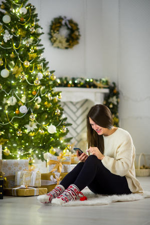 Christmas Eve. Cute dark-haired girl sits near the Christmas tree with the mobile phone in hands. House is decorated by holiday. Under Christmas tree a lot of beautifully wrapped up boxes with gifts.の写真素材