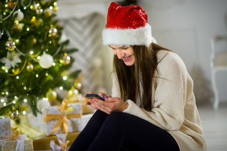 Christmas Eve. Nice girl in Santa's cap sits near Christmas tree with the mobile phone in hands. House is decorated by holiday. Under Christmas tree there are a lot of gifts. Girl joyfully smiles.の写真素材
