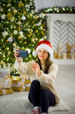 Christmas. Lovely girl in Santa's cap makes a selfie against the background of the Christmas tree. Under an elegant Christmas tree there are a lot of beautiful boxes with presents.の写真素材