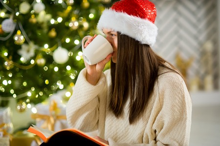 Girl in Santa's cap drinks coffee near the Christmas tree. Girl has long dark hair. Before her open notebook. Girl has thought. Christmas tree shines sparks.の写真素材