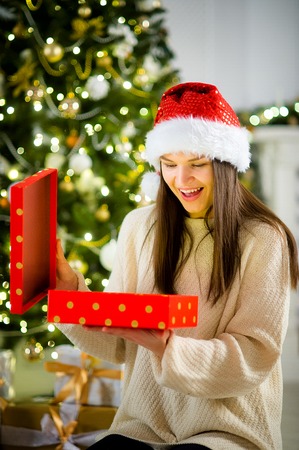 Boxing day. Nice girl the brunette opening a box with the Christmas present. On her face joy. Behind the girl beautifully decorated Christmas tree.の写真素材