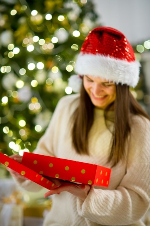 Boxing day. Nice girl the brunette opening a box with the Christmas present. On her face joy. Behind the girl beautifully decorated Christmas tree.の写真素材