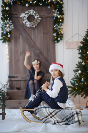 Two children near wooden door decorated with garlands and a wreath. Girl sits on steps and waves a hand. Boy in Santa's cap sits on sledge covered with a plaid. One can see the Christmas tree branchesの写真素材