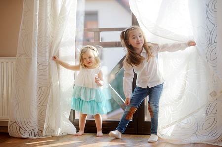 Two little sisters play in the room. Girls hide behind curtains, and then joyfully jump out because of curtains.の写真素材