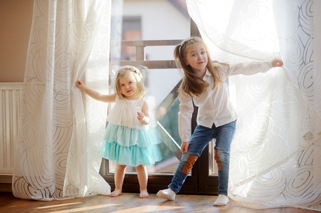 Two little sisters play in the room. Girls hide behind curtains, and then joyfully jump out because of curtains.