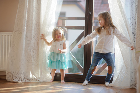 Two little sisters play in the room. Girls hide behind curtains, and then joyfully jump out because of curtains.の写真素材