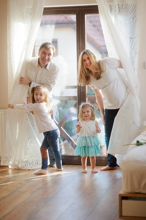 Cheerful family of four people. Two little sisters move apart curtains behind which parents hide. Young man and his pregnant wife play with daughters.の写真素材