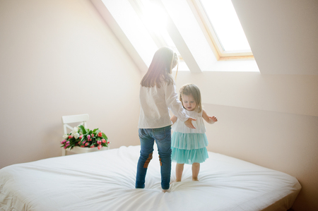 Two little sisters dance on a big bed. To girls it is very cheerful. On a white chair the big beautiful bouquet of tulips lies. From a window the sunlight flows.の写真素材