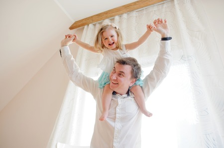 The young man gaily spends time with the little daughter. Charming baby sits on shoulders at the father and cheerfully laughs.の写真素材