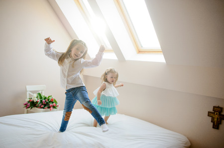 Two little sisters dance on a big bed. To girls it is very cheerful. On a white chair the big beautiful bouquet of tulips lies. From a window the sunlight flows.の写真素材