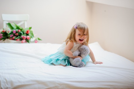 The charming little girl sits on a big bed. In hands at the baby a gray teddy bear. The girl has a cheerful playful look and the hair tousled from a play.の写真素材