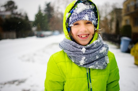 Portrait of the schoolboy walking in winter day. The boy is dressed in a bright jacket and a knitted cap and a scarf. His face and clothes are closed up with snow. The child happily smiles.の写真素材