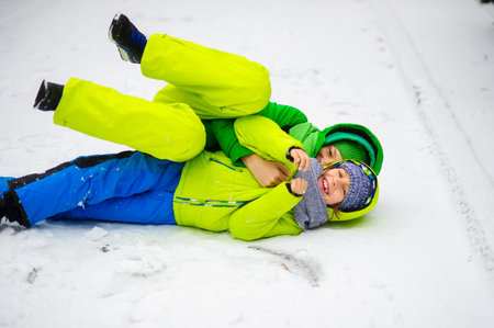 Two boys in bright winter overalls play on snow. It is snowing. Boys like to roll on a snow. They...