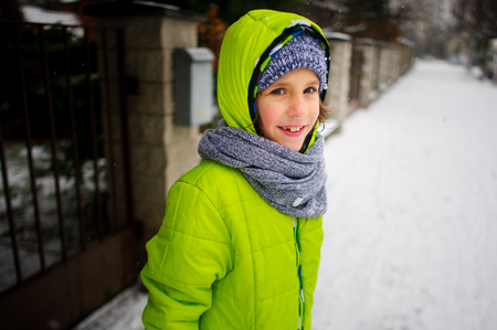 Little schoolboy goes down the street in winter day. The boy is dressed in a bright ski suit. It is snowing. The child likes such weather. He cheerfully smiles.の写真素材