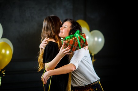 The young woman congratulates the girlfriend and gives her a box with a gift. Women joyfully embrace. The room is decorated with balloons.の写真素材