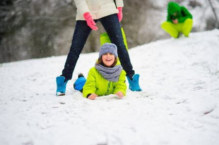 Cheerful boy of school age cheerfully spends time in winter day. Boy in a bright ski suit slides on snow lying on a stomach. He slips between mother's legs. To boy it is very cheerful.の写真素材
