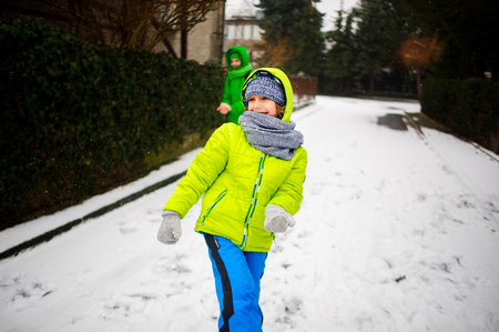 Two boys cheerfully play on the street in winter day. It is snowing. Boys are dressed in warm bright ski suits. They rejoice to the first snow.の写真素材