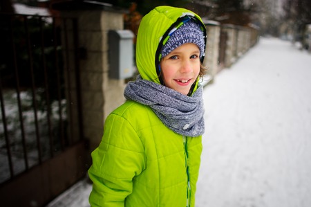 Little schoolboy goes down the street in winter day. The boy is dressed in a bright ski suit. It is snowing. The child likes such weather. He cheerfully smiles.の写真素材