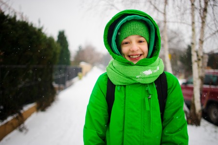 The cheerful teenager goes down the street in winter day. Behind shoulders at him a backpack. The boy is dressed in a bright green ski suit. It is snowing. The fellow smiles from pleasure.の写真素材