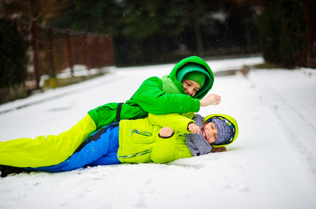 Two boys in bright winter overalls play on snow. It is snowing. Boys like to roll on a snow. They joyfully laugh.の写真素材
