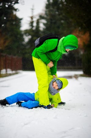 Two boys in bright winter overalls play on snow. The older boy helps to stand up younger. It is snowing. Children cheerfully laugh.の写真素材
