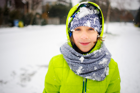 Portrait of the schoolboy walking in winter day. The boy is dressed in a bright jacket and a knitted cap and a scarf. His face and clothes are closed up with snow. The child happily smiles.の写真素材