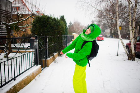 The cheerful teenager goes down the street in winter day. Behind shoulders at him a backpack. The boy is dressed in a bright green ski suit. It is snowing. The fellow smiles from pleasure.の写真素材