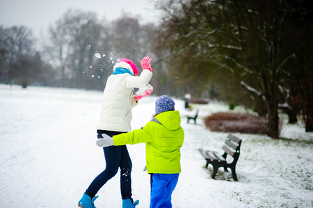 Young woman and her son cheerfully spend time in winter park. They have started battle by snowballs. And though it is little snow, mother and son are very glad. Both are dressed in bright ski suits.の写真素材