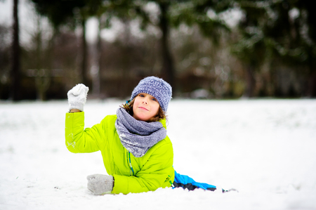 The boy of school age plays snowballs in the winter park. He is dressed in warm and bright winter overalls. The fellow lies on the ground covered with snow and throws a snowball. To him it is very cheerful.の写真素材