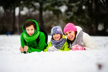 Charming young woman and two of her sons cheerfully spend time in the winter park. They are dressed in bright warm ski suits. All look out because of a big snowdrift.の写真素材