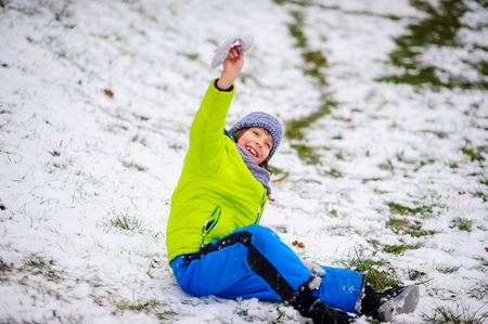 The boy of school age in bright winter overalls sits on the ground which is slightly covered with the first snow. Through snow the grass is visible. The boy rejoices. Children adore winter and snow.の写真素材