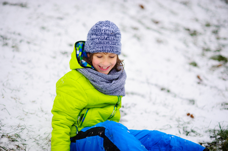 The boy of school age in bright winter overalls sits on the ground which is slightly covered with the first snow. Through snow the grass is visible. The boy rejoices. Children adore winter and snow.の写真素材