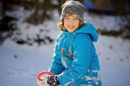 Winter portrait of the boy of younger school age. The nice fellow in a blue jacket and a gray knitted cap joyfully smiles. He rejoices to snow. Snow on clothes and the boy's hair.の写真素材