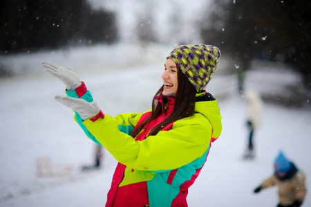 The young woman joyfully holds up palms under the falling snow. She on walk in the winter park. The woman is dressed in a bright jacket and knitted gloves and a cap. The falling snow causes delight.の写真素材