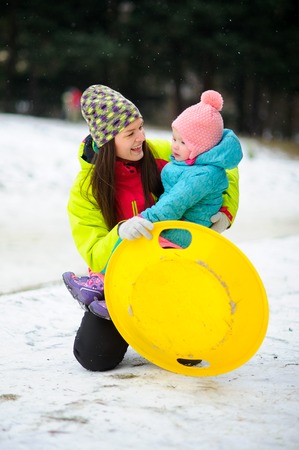 Mother with little daughter have come to ride the sledge to winter park. Both are dressed in bright warm ski suits. Woman holds the yellow saucer sledge in hand. It is snowing.の写真素材
