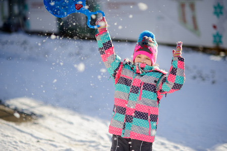 Girl of younger school age on walk in winter day. Child warmly is also brightly dressed. In hands at the girl the light-weight saucer sledge for driving from an ice slope.の写真素材
