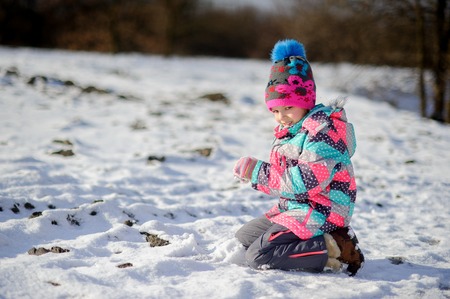 Girl of younger school age in bright ski suit made a snowball. She is kneeling the ground which is slightly covered with snow. Children very much love winter and snow.の写真素材