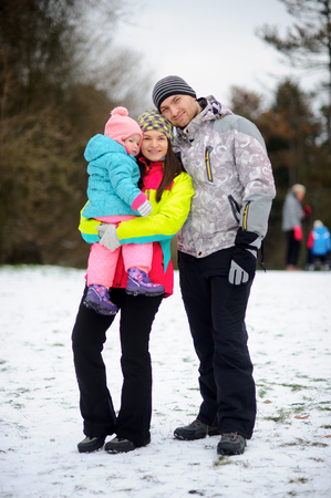 Winter family portrait. Young couple with little daughter against the background of the winter park. Family stands having embraced. Man and woman smile. Baby has a serious look.の写真素材