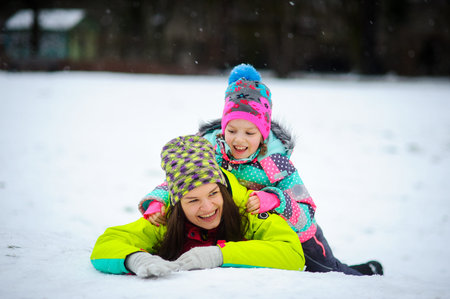Lovely young woman and girl in bright jackets lie on snow. It is snowing. Mother and the daughter...