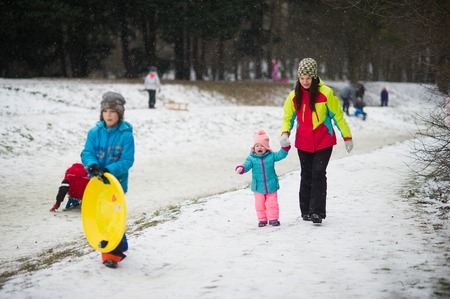 Young mother has brought little daughter into winter park. Both are dressed in bright warm ski suits. In park there are a lot of people riding the sledge. It is snowing. Baby for some reason cries.の写真素材