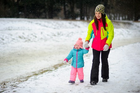 Young mother has brought the little daughter into the winter park. Both are dressed in bright warm ski suits. The woman holds the baby by a hand. It is snowing. Baby for some reason cries.の写真素材