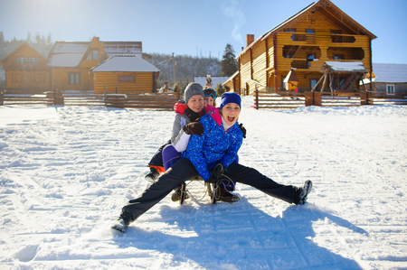 Cheerful driving on the sledge. Two women and the child ride one sledge. Sunny winter day. Wooden cottages. Coniferous forest in the distance.の写真素材
