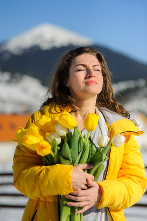 Charming woman with a bouquet of tulips against the background of snow-covered mountain tops. She is dressed in a bright yellow down-padded coat. Woman has a happy look.の写真素材